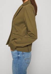 Žena oblečená v olivově zeleném blejzru s kapsičkami a modrých džínách, stojící v profilu s rukama v předních kapsách.