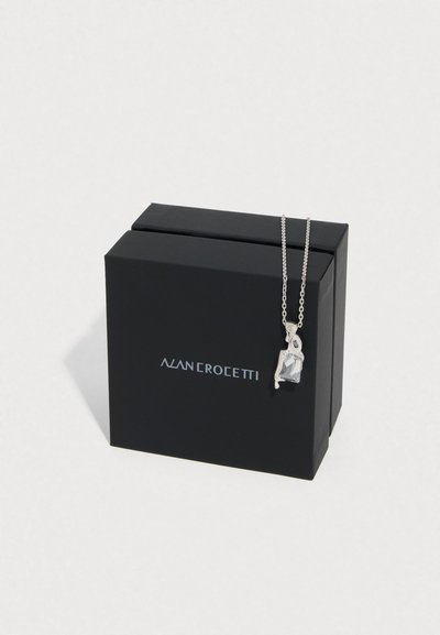 Hõbedast ripats-kaelakee lõigatava kristallikiviga, eksponeeritud musta ehtekarbi kõrval, millel on valge bränding.