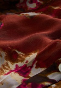 Stoff mit einer welligen Textur auf einem satten rotbraunen Grund, akzentuiert durch florale Muster in Rosa- und Cremetönen. Weiches Material.