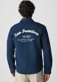 Camisa de manga larga azul marino con puños abotonados, que presenta texto bordado en la parte posterior: "San Francisco" y "Colección de Artesanía."