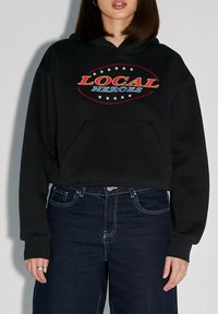 Czarna krótka bluza z kapturem z przednią kieszenią kangurką, z napisem "LOCAL HEROES" w czerwonym i niebieskim, otoczonym białymi gwiazdkami na klatce piersiowej.