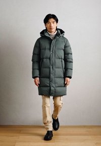 Tummanvihreä puhvitakki, jossa on huppu, tikatut osat, nappikiinnitys ja sivutaskut, päällä vaaleat housut ja mustat kengät.