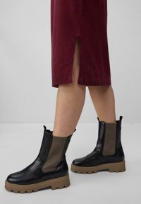 Schwarze Stiefeletten aus Leder mit strukturierten Netzpaneelen, massiven braunen Gummisohlen und Ziehschlaufen. Getragen mit einem weinroten Kleid, das seitliche Schlitze aufweist.