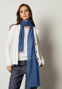 Blå uldscarf med en glat tekstur, prydet med frynser, lagdelt over en hvid top og en cremefarvet cardigan med ribbet mønster og knapper.