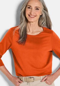 Orange Langarmoberteil mit rundem Ausschnitt und glatter Oberfläche, kombiniert mit einem hellen Gürtel und beigen Hosen.
