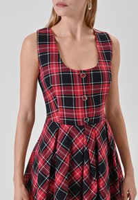 Abito a quadri rosso e nero con scollo rotondo, corpetto strutturato e gonna plissè. Presenta tre bottoni circolari sul davanti.