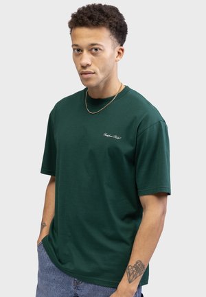 Crew-neck, short-sleeve T-shirt in dark green cotton. Features small white embroidered logo on chest. Model wears blue denim shorts.