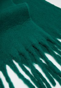 Bufanda de lana verde azulado con una superficie suave y texturizada, que presenta extremos con flecos y una forma ancha y rectangular.