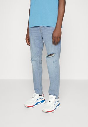 Homme portant un jean bleu clair déchiré, des baskets blanches avec des accents rouges et bleus, et une chemise bleue.