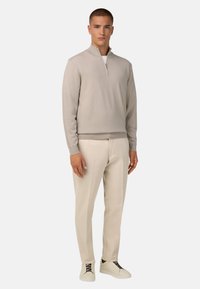 Béžový pletený svetr s polovičním zipem, ribbenými manžetami a spodním lemem, v kombinaci s světle béžovými kalhotami a bílými teniskami s černými tkaničkami.