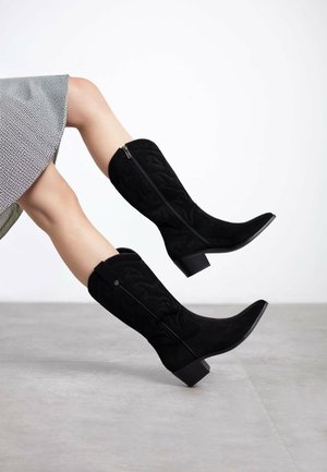 Botas negras de ante hasta la rodilla con un patrón texturizado, punta afilada y tacón grueso; cuentan con una cremallera lateral y un acento decorativo en la parte posterior.