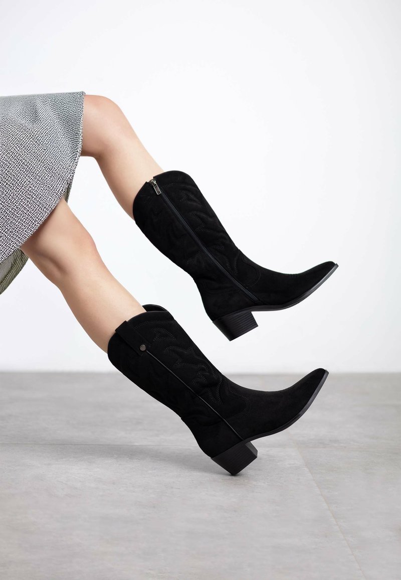 Botas negras de ante hasta la rodilla con un patrón texturizado, punta afilada y tacón grueso; cuentan con una cremallera lateral y un acento decorativo en la parte posterior.