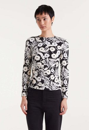 Joven con pelo negro corto, vistiendo una blusa de manga larga con un patrón floral en blanco y negro y pantalones negros, de pie contra un fondo liso.