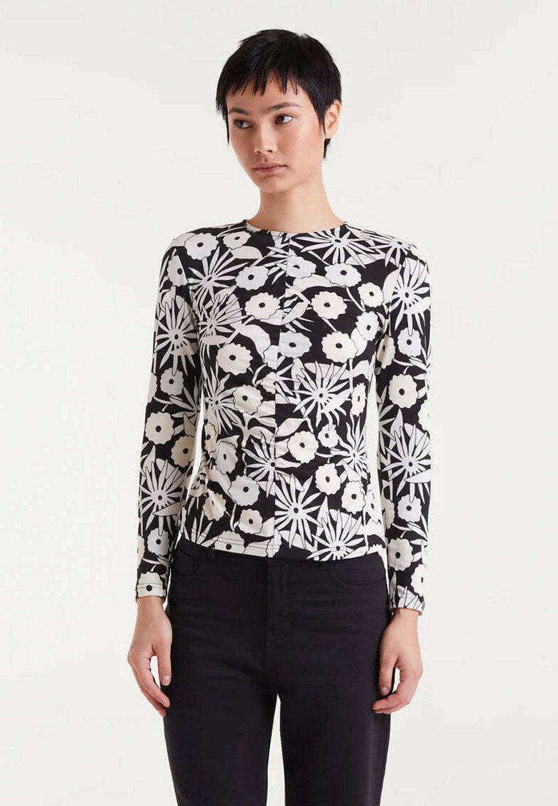 Joven con pelo negro corto, vistiendo una blusa de manga larga con un patrón floral en blanco y negro y pantalones negros, de pie contra un fondo liso.