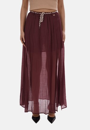 Donna che indossa una lunga gonna bordeaux trasparente con cintura bianca a corda e scarpe nere a punta, in piedi davanti a uno sfondo bianco.