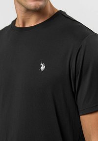 Černé bavlněné tričko s kulatým výstřihem. Obsahuje malé bílé logo na hrudi. Hladká textura s krátkými rukávy.