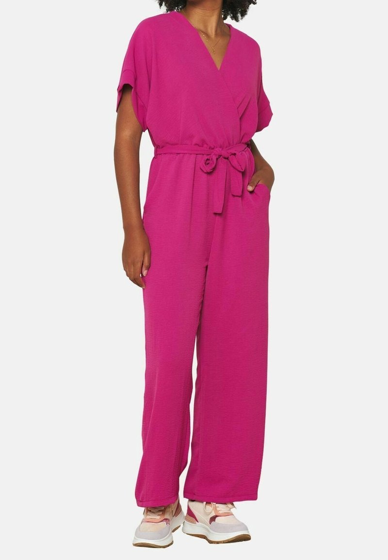 Personne portant une combinaison rose vif à manches courtes avec une ceinture nouée à la taille et des baskets blanches et roses, se tenant debout avec une main dans la poche.