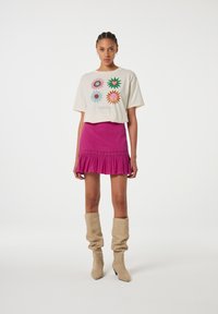 Crème graphic t-shirt met kleurrijke bloemenprints, gecombineerd met een roze corduroy rok met ruches aan de onderkant, en beige slouchy kniehoge laarzen.