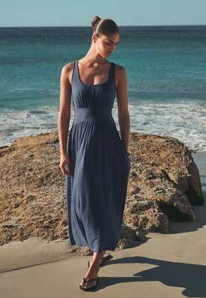 Nő ujjatlan sötétkék maxiruhában és szandálban sétál homokos tengerparton nagy szikla mellett, az óceán hullámai a háttérben.