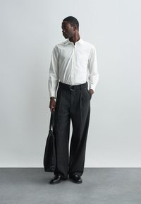 Bijela košulja dugih rukava, crne široke hlače sa prugama, crni pojas i kožne cipele. Crna torba prebačena preko ramena na neutralnoj pozadini.