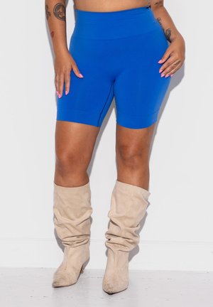 Rond persoon met felblauwe fietsshorts en beige slappe laarzen, staand tegen een effen witte achtergrond.