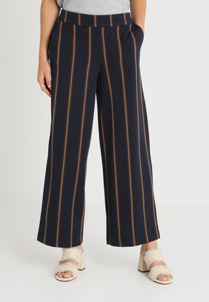 Persona que lleva pantalones anchos azul marino con rayas verticales marrones y sandalias beige texturizadas de tacón ancho y punta abierta, de pie con las manos en los bolsillos.