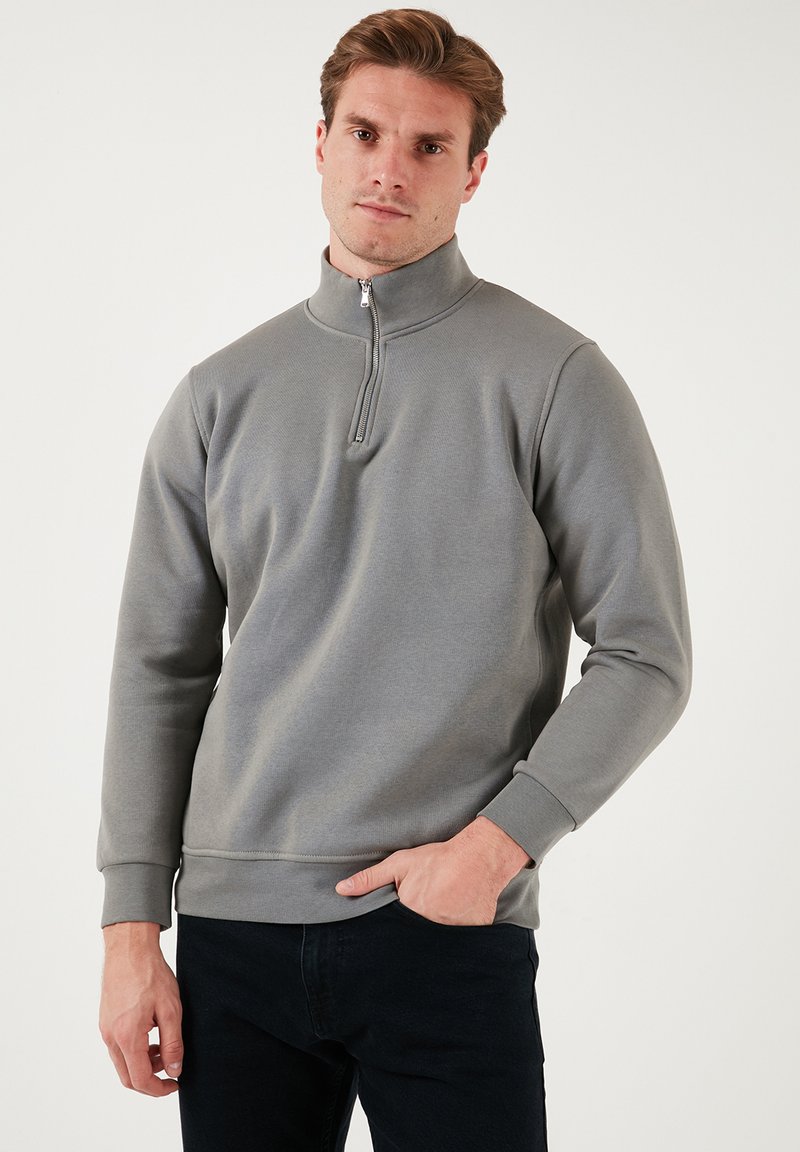 Felpa zip in grigio chiaro con colletto alto e polsini a costine, caratterizzata da una trama liscia e una vestibilità casual, abbinata a jeans scuri.