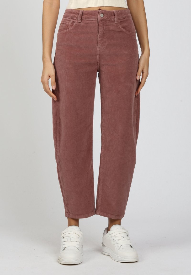 Nainen, joka pukeutunut vaaleanpunaisen pörröiseen korkevyötäröisiin suoralahkeisiin corduroy-housuihin ja valkoisiin lenkkareihin, seisoo yksinkertaisen taustan edessä.