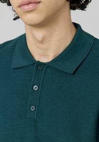 Polo en teal fait en tissu texturé, avec un col et trois boutons. La vue rapprochée met en avant les détails de l'encolure et des boutons.