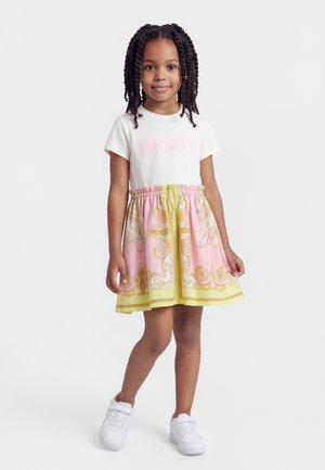 Young girl with braided hair wearing a white Versace t-shirt, pink and yellow patterned skirt, and white sneakers, standing on a white background.