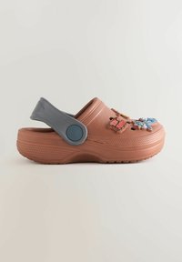 Sabots en caoutchouc marron avec une sangle arrière réglable grise et des charms décoratifs en plastique sur le dessus, dotés de trous perforés pour la respirabilité.