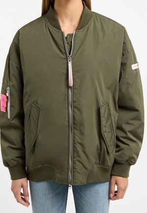 Veste bomber verte zippée avec col, poignets et bas côtelés, portée sur un jean bleu, avec des poches et des patches roses sur les manches.