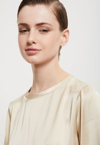 Krémová saténová blúzka s krátkymi rukávmi a okrúhlym výstrihom, s jemným leskom a subtílnym drapovaním pre hladkú textúru.