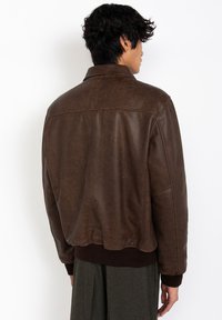 Chaqueta de cuero marrón con una textura ligeramente brillante, que presenta un cuello, dobladillo acanalado y costuras en los hombros. La tela es suave y flexible.