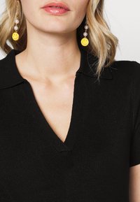 Top de malha ribada preta com decote em V e mangas curtas. Brincos com rostos sorridentes amarelos, adornados com pérolas, pendem de um suporte em tom dourado.