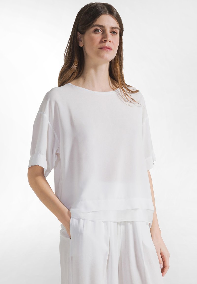 Vrouw met stijlbruin haar, gekleed in een los wit kortärmelig shirt en witte broek, staand met één hand in de zak tegen een witte achtergrond.