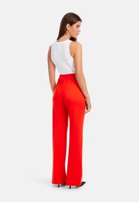 Rote weit geschnittene Hose mit hoher Taille und strukturierter weißer ärmelloser Bluse. Modell von hinten gesehen, auf schwarzen Absatzschuhen stehend.