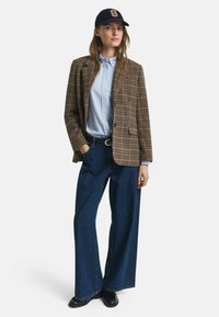 Donna che indossa un blazer a quadri marroni, una camicia azzurra con bottoni, jeans larghi neri e scarpe nere, oltre a un cappellino da baseball blu navy con una "G" arancione.
