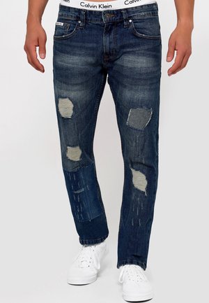 Jean déchiré bleu foncé avec patchwork porté par une personne en baskets blanches et taille visible Calvin Klein.
