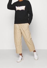 Sweat-shirt noir avec un logo graphique coloré, porté avec un pantalon cargo beige ample et des baskets blanches avec des accents noirs.