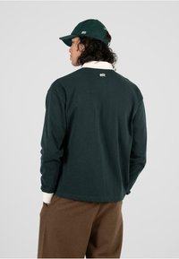 Camisa de manga larga verde oscuro con cuello y puños blancos; corte holgado; detalle del logo en la parte posterior; combinada con pantalones marrón claro.