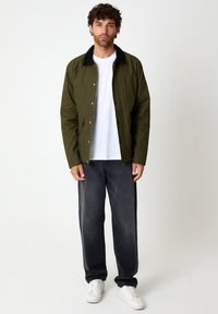 Giacca verde oliva con colletto nero, bottoni a pressione e tasche laterali, abbinata a jeans scuri e sneakers bianche. Texture del tessuto liscia.