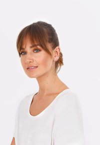 Modelo con una blusa blanca de manga corta y escote amplio. Lleva pendientes de botón rosas y tiene el cabello castaño liso con flequillo.