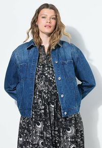 Denimjacka i mörkblått, med knäppning framtill, två bröstfickor och klassisk krage, bärs över en svart klänning med vita himmelska mönster.