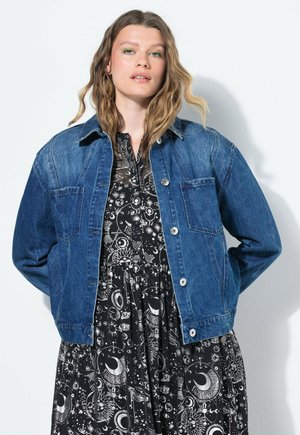 Chaqueta de mezclilla en azul oscuro, con cierre de botones, dos bolsillos en el pecho y un cuello clásico, llevada sobre un vestido negro con patrones celestiales en blanco.