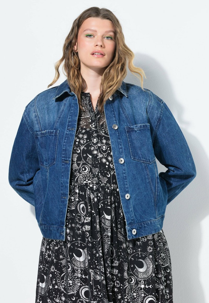 Denimjacka i mörkblått, med knäppning framtill, två bröstfickor och klassisk krage, bärs över en svart klänning med vita himmelska mönster.