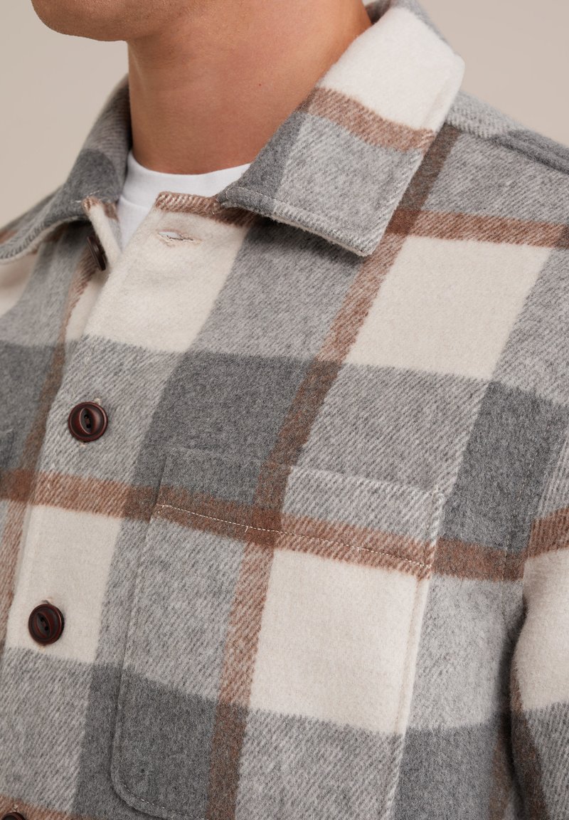 Kurtka w kratę z wełny w odcieniach szarości i kremu, z brązowymi akcentami, kołnierzem oraz kieszenią na piersi z ciemnymi guzikami. Miękka tekstura.