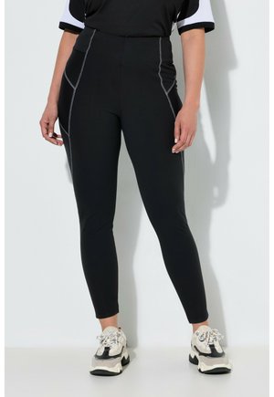 Kvinde iført sorte højttaljede leggings med kontrastsyning og hvide og sorte chunky sneakers, stående mod en lys væg.