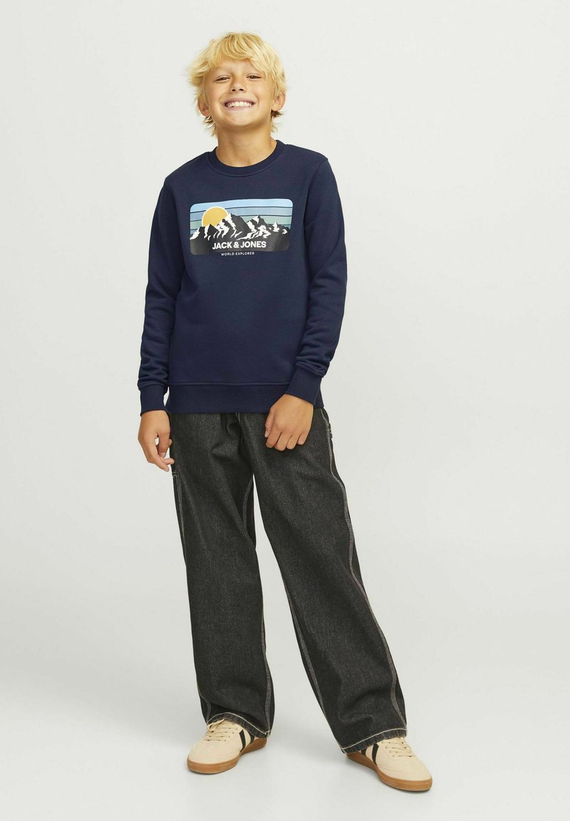 Navy blue sweatshirt with mountain graphic and text, paired with loose-fitting dark jeans and light beige sneakers. Casual outfit for youth.