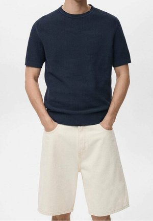 Homme portant un pull à manches courtes texturé bleu marine foncé et un short beige clair longueur genou, les mains dans les poches.